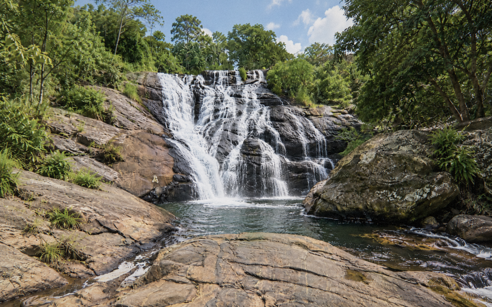 Água descendo por rochas em uma cachoeira cercada de árvores, com uma piscina natural e pedras em primeiro plano